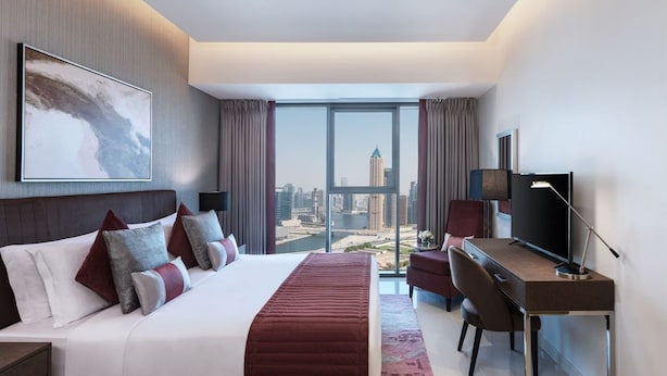 A modern hotel room with a large bed, a desk, a TV, and a city view through large windows. - Damac Maison Aykon City Dubai (Dubai)