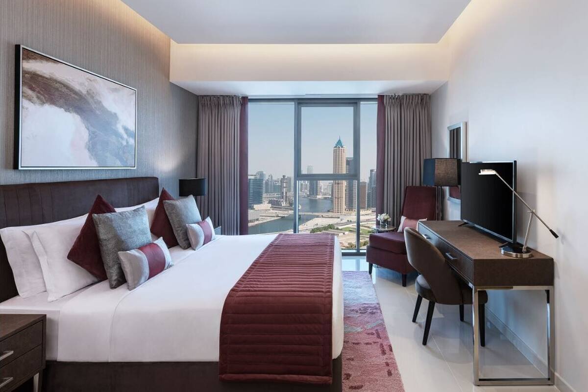 A modern hotel room with a large bed, a desk, a TV, and a city view through large windows.