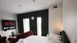 A modern hotel room with a large bed, a desk, a chair, a TV, and a window with blinds. - Affordable Classy Studio in Downtown Des Moines! (Des Moines)