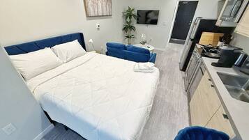 Una habitación de hotel moderna con una cama grande, un pequeño espacio para sentarse, una kitchenette y un televisor fijado a la pared.