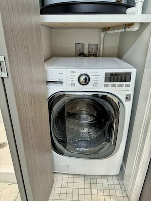 A compact, modern washing machine with a digital display, located in a small laundry room with tiled flooring.