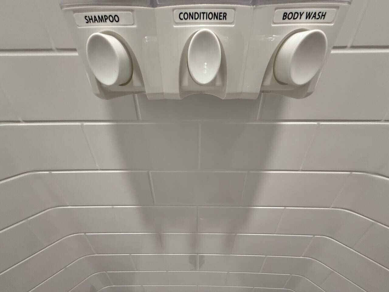 Casa de banho com três dispensadores rotulados 'SHAMPOO', 'CONDICIONADOR' e 'ALMOFADA DE CORPO'.