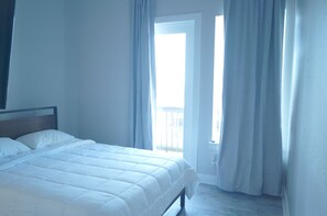 A bedroom with a bed, a window with curtains, and a door leading to an outdoor area.