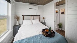 Un dormitorio con techo de madera, una cama con ropa blanca, una mesita de noche con una planta y una ventana con vistas al exterior.