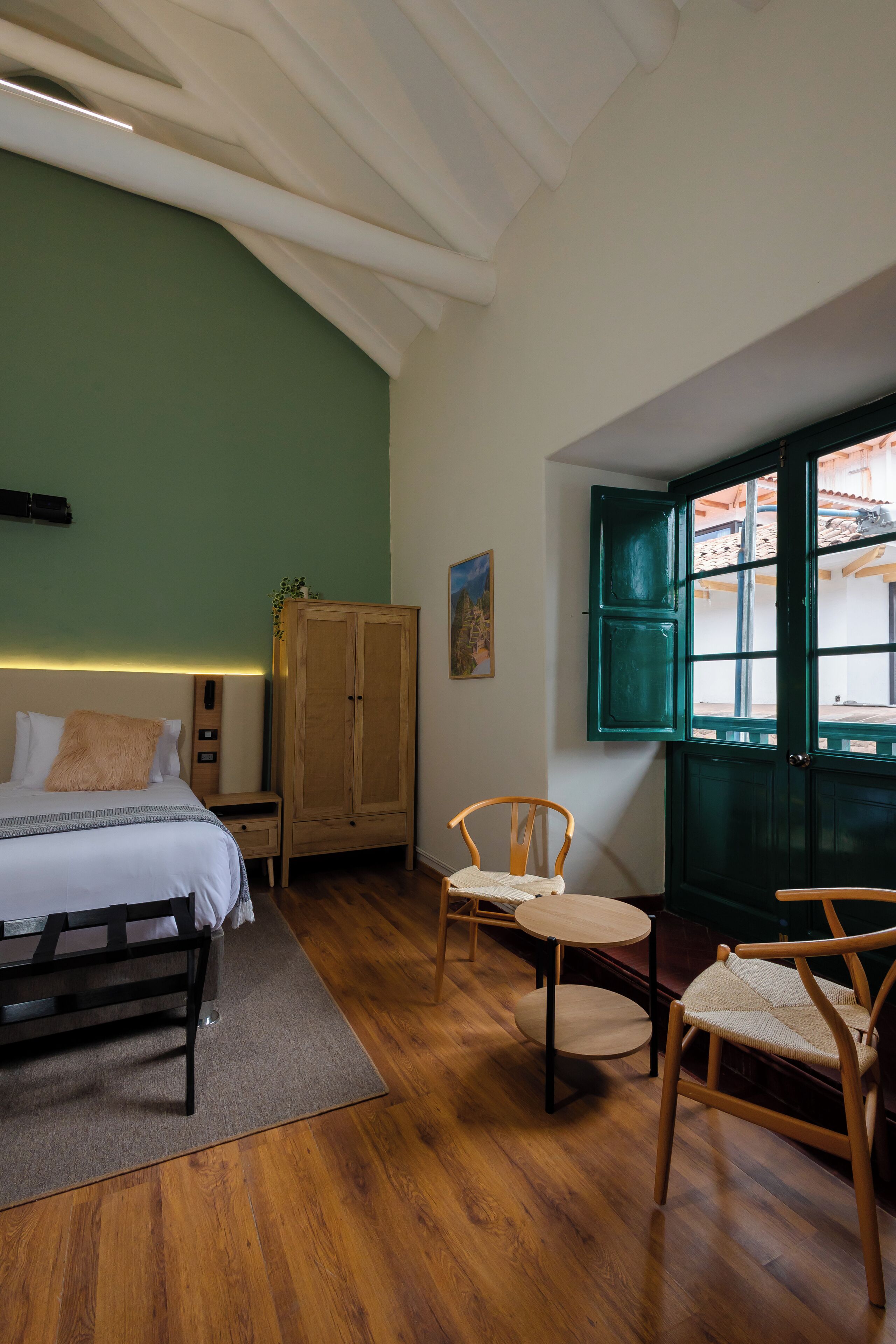 A bedroom with a bed, wooden wardrobe, two chairs, a small table, and a window with green shutters.