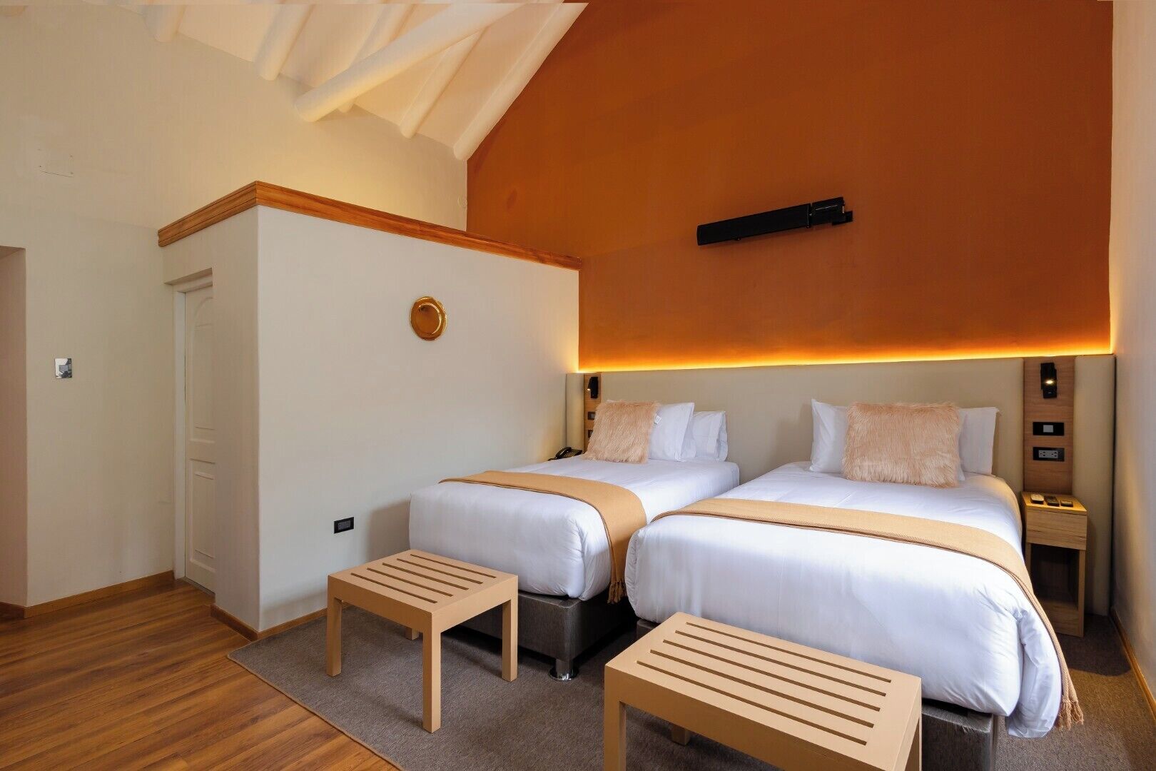 Habitación de hotel con dos camas, un banco de madera, una mesita pequeña y una lámpara en el techo.