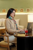 Een persoon zit aan een tafel met een laptop, een glas rode drank en een lamp.