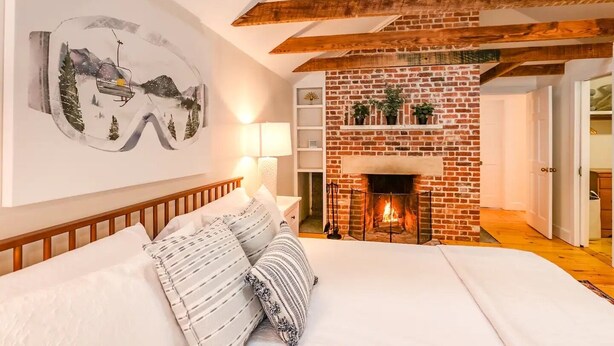 Een slaapkamer met een schouw van baksteen, houten balken, een bed met witte beddengoed en een aan de muur hangend schilderij.