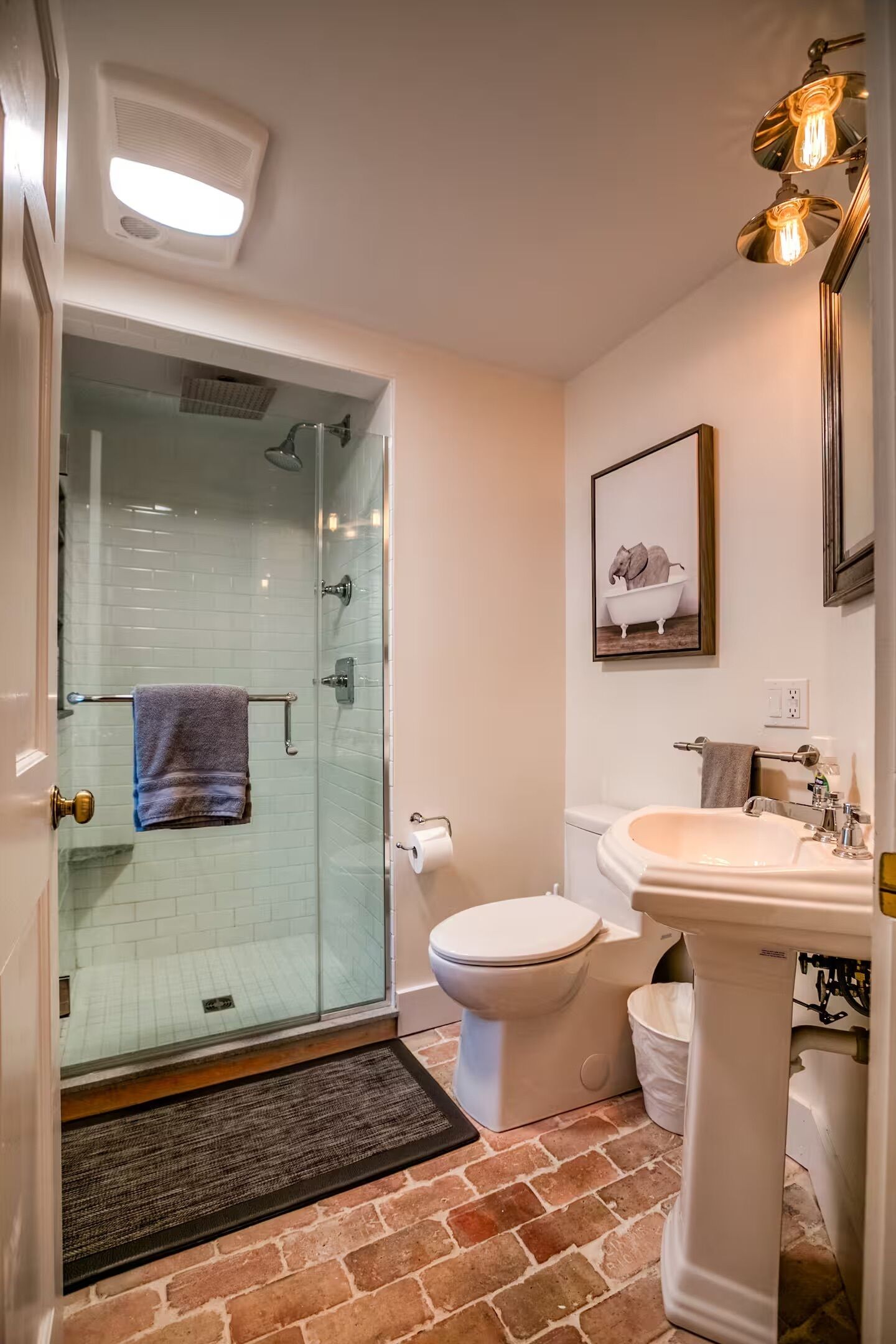 A bathroom with a glass shower enclosure, a white toilet, a pedestal sink, and a framed picture of an elephant in a bathtub.