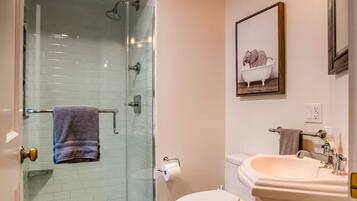 A bathroom with a glass shower enclosure, a white toilet, a pedestal sink, and a framed picture of an elephant in a bathtub.