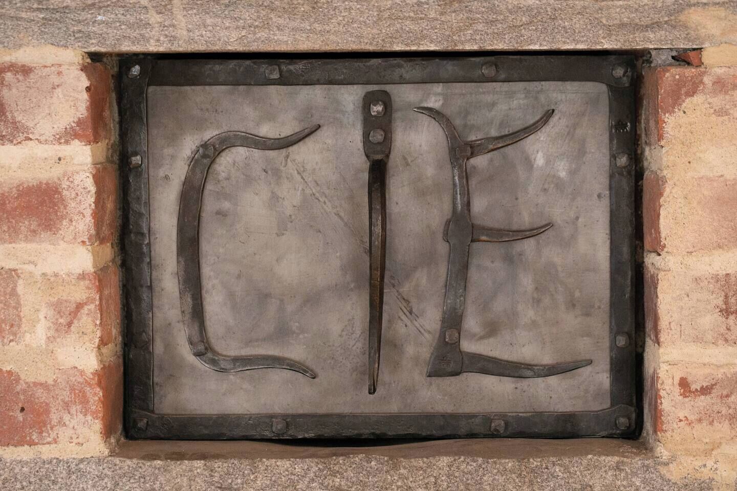 Une plaque métallique portant les lettres « C E » et une lame de couteau au centre, fixée à un mur de briques.