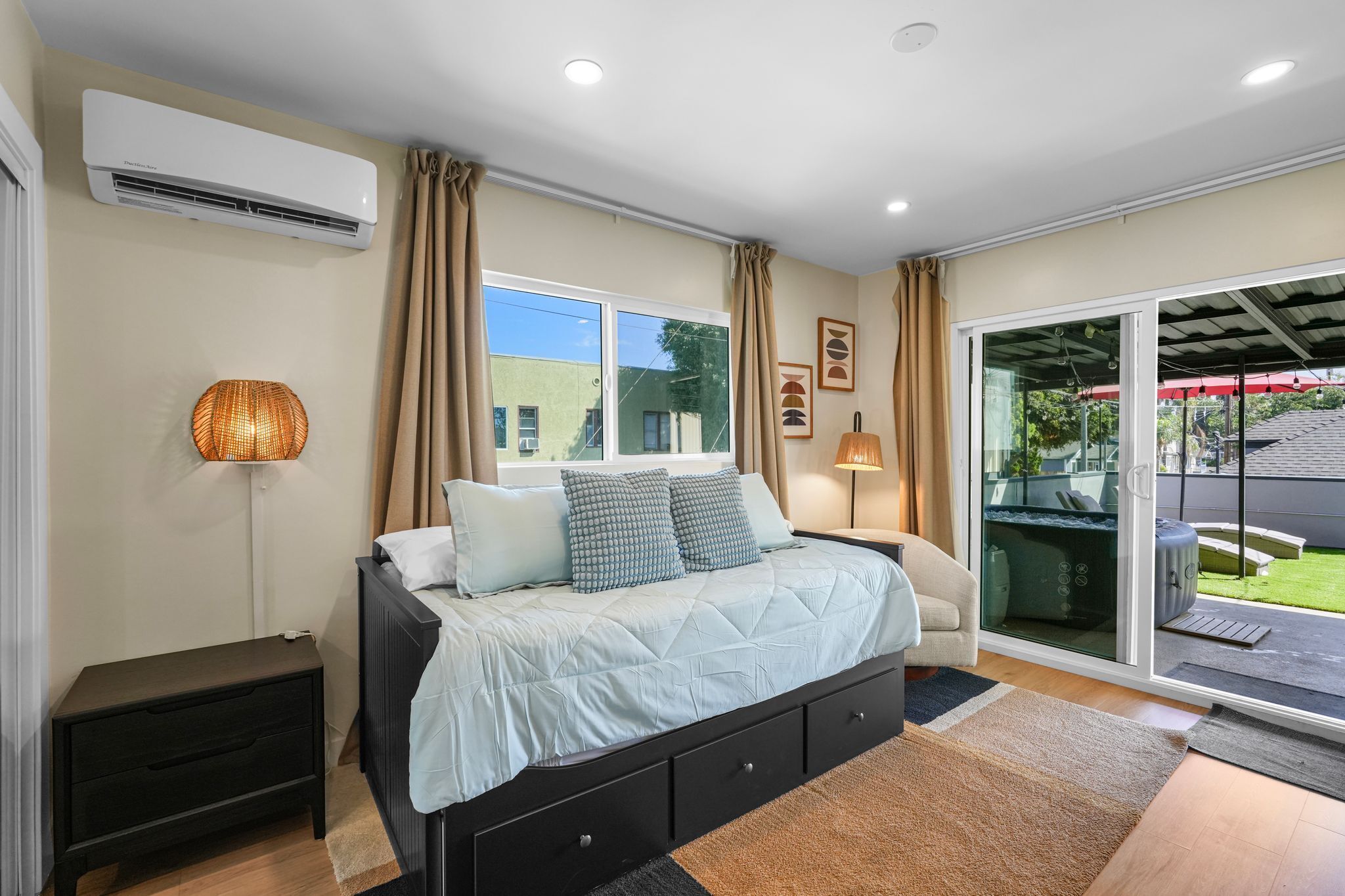 A bedroom with a bed, bedside tables, an air conditioner, a window with curtains, and a sliding glass door leading to an outdoor area.