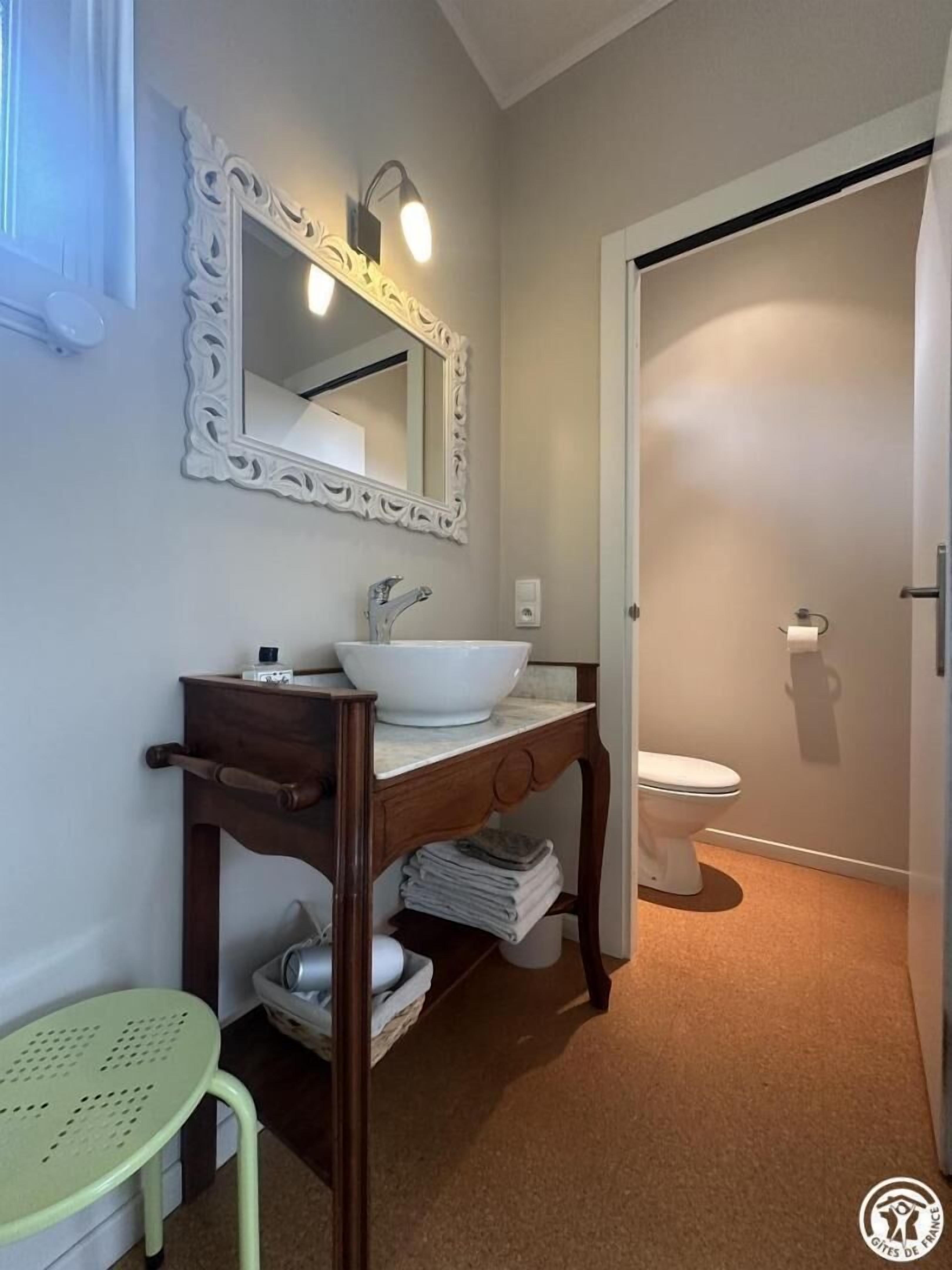 Une salle de bain avec un lavabo blanc, un miroir et des toilettes. On y trouve un meuble-lavabo en bois, un porte-serviettes et un petit tabouret vert.
