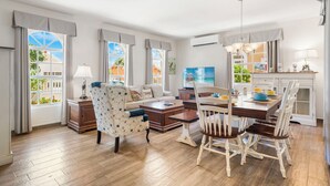 Ein modernes Wohnzimmer mit Sofa, Sessel, Esstisch und Stühlen. Es gibt Fenster mit Vorhängen, ein Bild an der Wand und einen Fernseher.