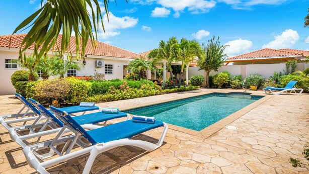 A pool area with lounge chairs, a swimming pool, and a building with a tiled roof.
