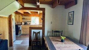 Ein Essbereich mit einem Holztisch, Stühlen und einer Blumenvase. Im Hintergrund ist eine Küche mit Holzschränken und einem Fenster erkennbar.