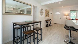 A modern kitchen with a wooden table and bar stools, a large mirror, and a dining area with a white table and bar stool.