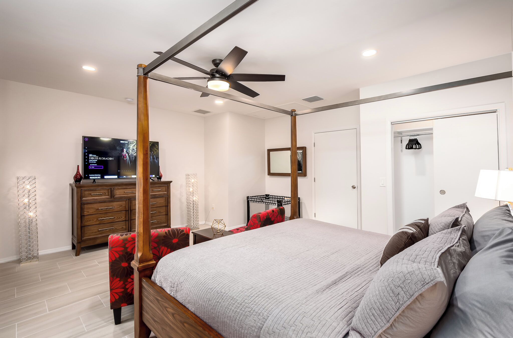 A modern bedroom with a four-poster bed, a ceiling fan, a flat-screen TV, a wooden dresser, and a wardrobe.