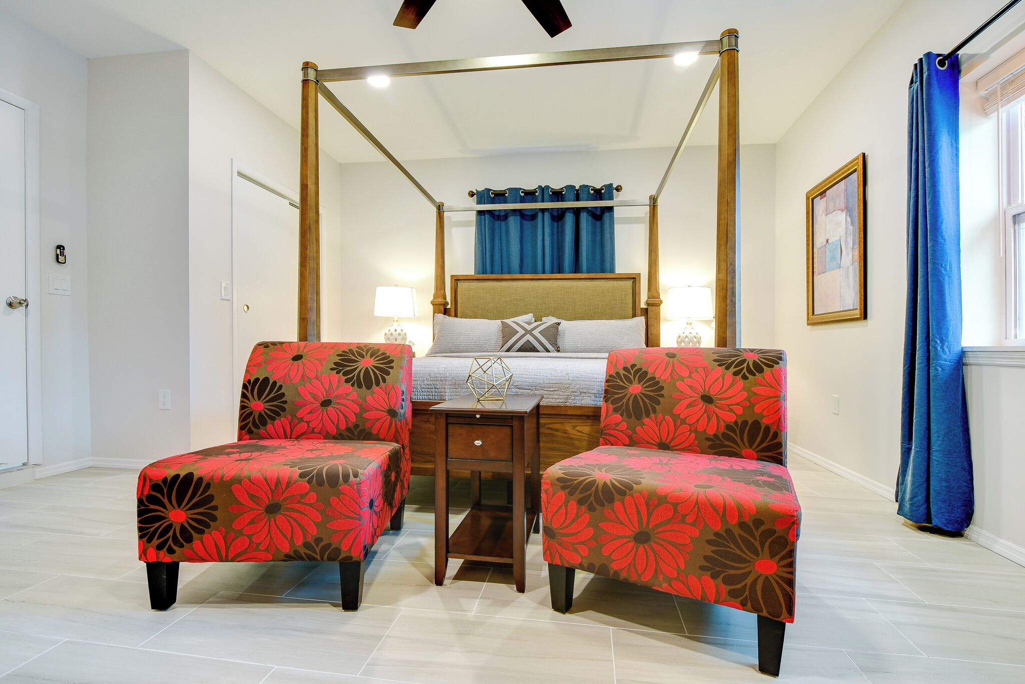A four-poster bed with a wooden frame and a canopy, flanked by two armchairs with floral upholstery.