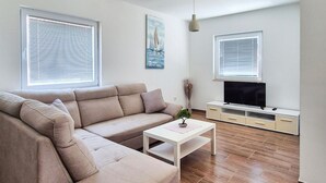 Un salon moderne avec un canapé beige, une table basse blanche, un téléviseur posé sur un meuble blanc et deux fenêtres équipées de stores.