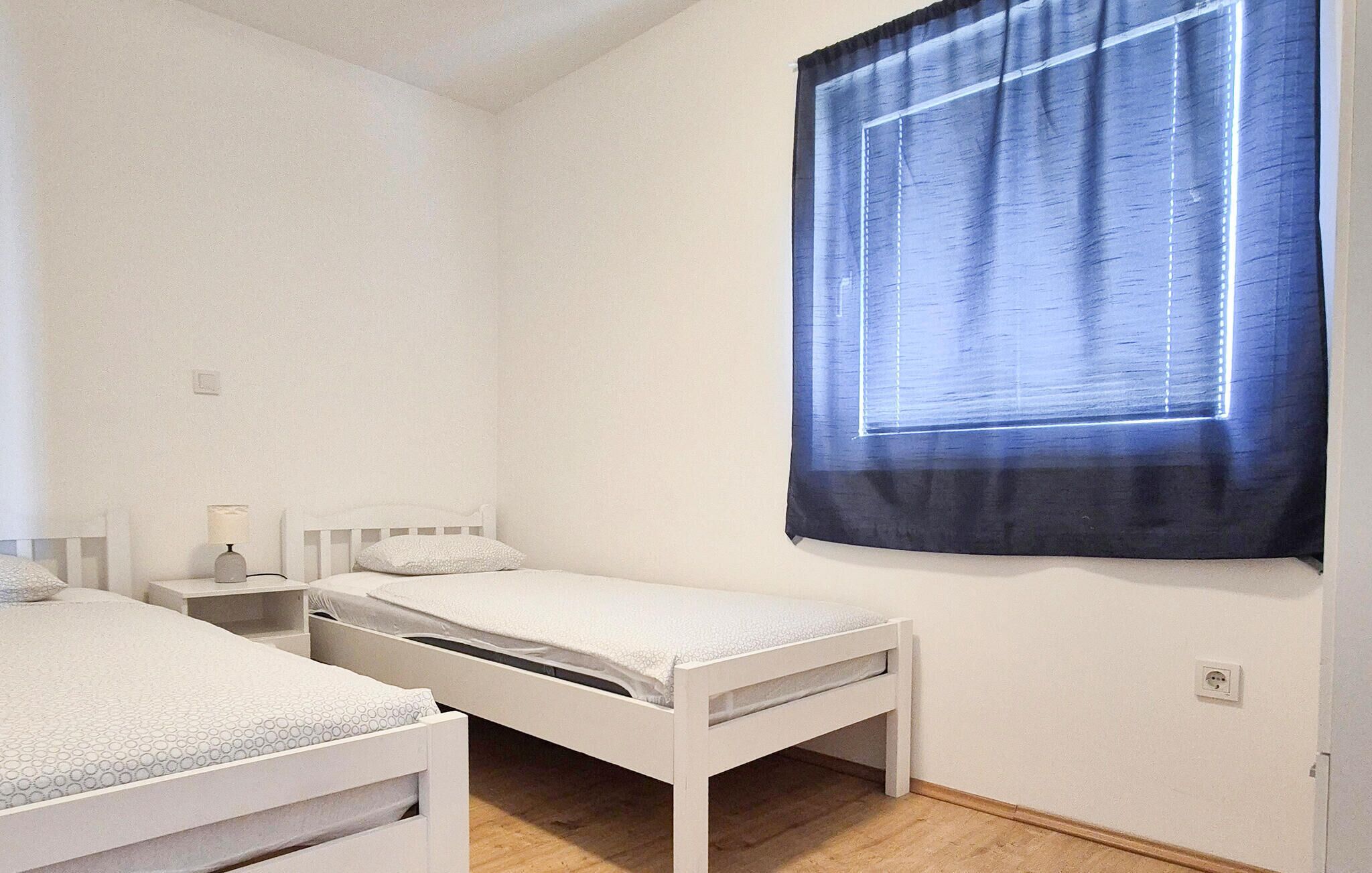 A small, clean room with two single beds, a bedside table with a lamp, and a window with a blue curtain.