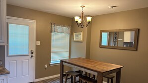 A dining area with a wooden table and chairs, a chandelier, a door with a welcome mat, and a window with blinds. - The Cottage at The Stables - Charming 1 bedroom - Sleeps 5 (Vancleave)