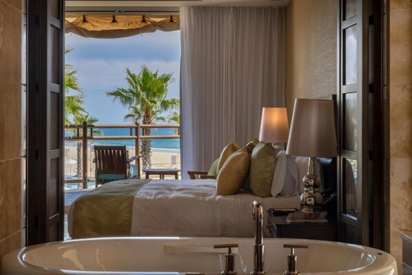 Ein Hotelzimmer mit einer großen Badewanne, einem Bett mit vielen Kissen, einem Nachttisch mit Lampe und Blick auf Strand und Palmen.