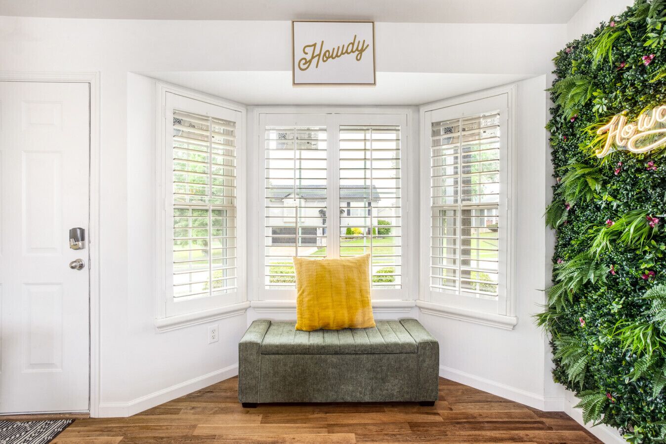 A room with a bay window, a green sofa with a yellow pillow, and a wall-mounted green plant arrangement with a neon sign.