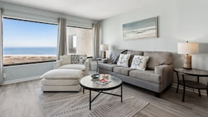 Ein modernes Wohnzimmer mit Blick auf den Strand, ein graues Sofa, ein weißer Sessel und ein runder Couchtisch.