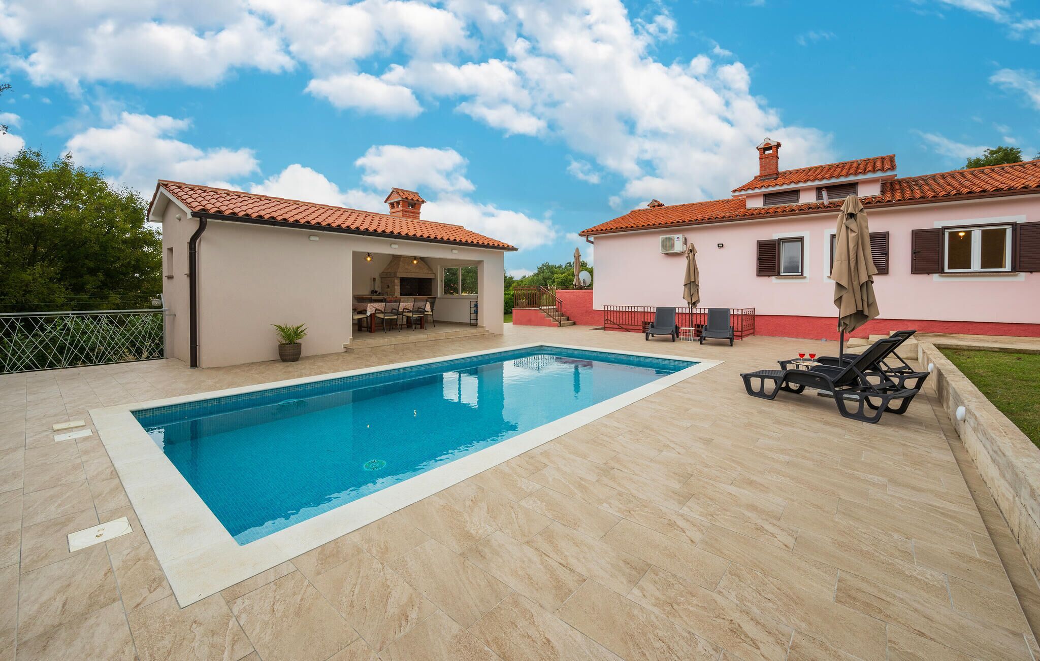 A swimming pool with a tiled deck, a covered patio area with outdoor furniture, and a building with a red-tiled roof.