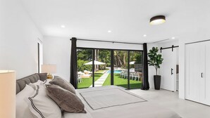 A modern bedroom with a large bed, a view of a backyard with a pool, and a sliding door leading to a patio area. - Villa Parque Pickleball/Volleyball/Bball/MiniGolf (Miami)