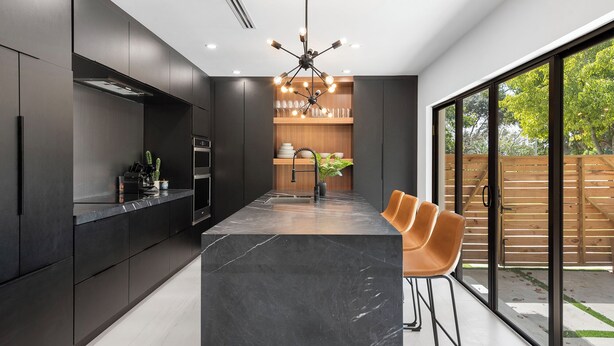 A modern kitchen with dark cabinetry, a marble island, and a large window offering a view of the outdoors. - Villa Parque Pickleball/Volleyball/Bball/MiniGolf (Miami)
