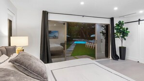 A modern bedroom with a large bed, a bedside table with a lamp, a sliding glass door leading to a pool area, and a potted plant. - Villa Parque Pickleball/Volleyball/Bball/MiniGolf (Miami)