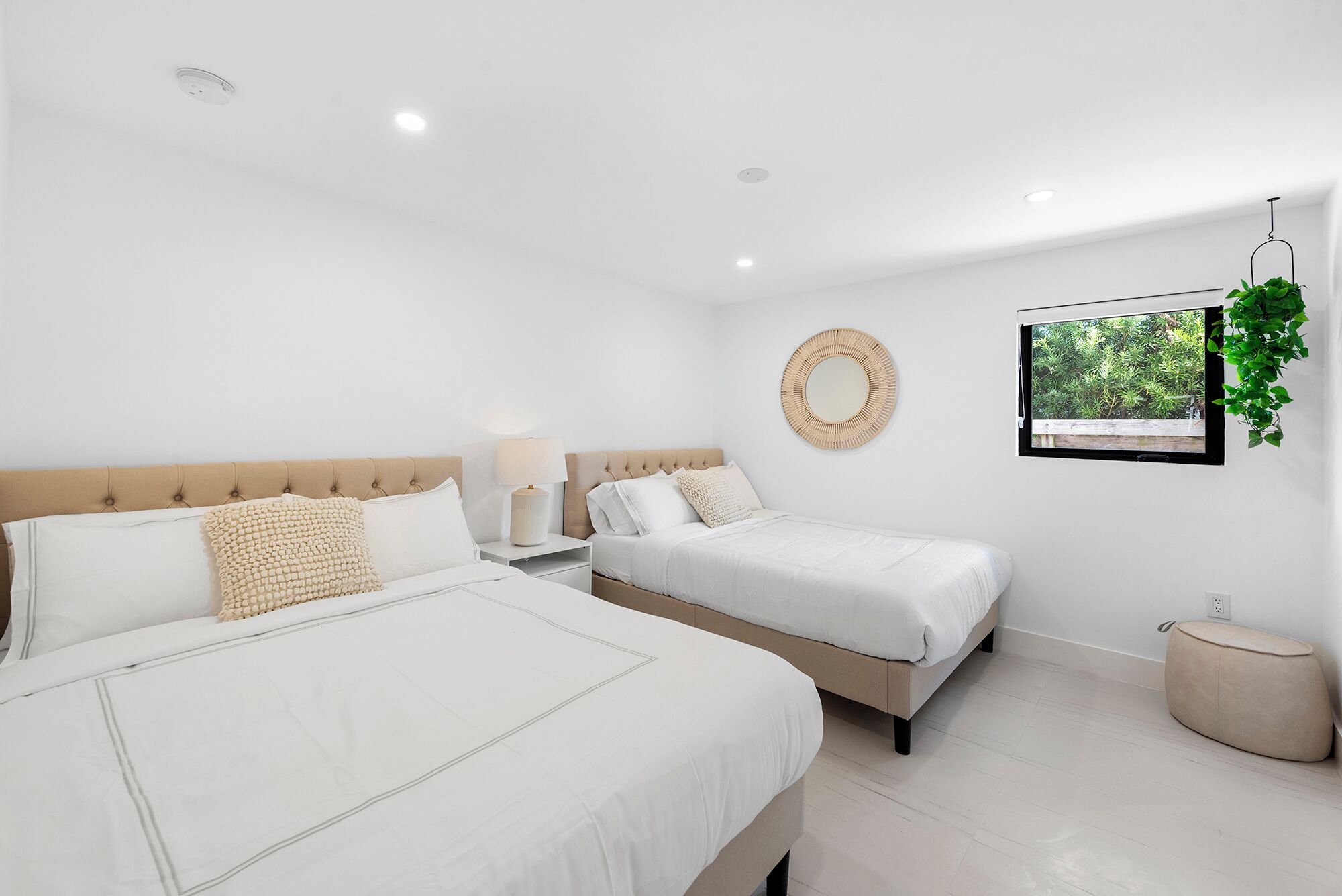 Quarto moderno com duas camas, um espelho redondo, mesa de cabeceira com abajur e janela com vista para o verde.