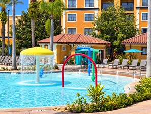 A swimming pool with a water play area, surrounded by lounge chairs and a building with balconies.