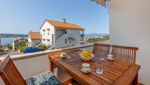 Un balcon avec une table en bois dressée pour deux personnes, un panier de fruits et une vue sur la mer et les bâtiments.