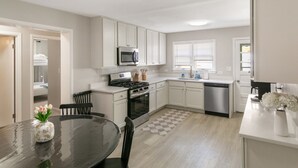 Cocina moderna con armarios blancos, estufa de acero inoxidable, microondas y lavavajillas. Hay un comedor con mesa y sillas, y un jarrón con flores sobre la mesa.