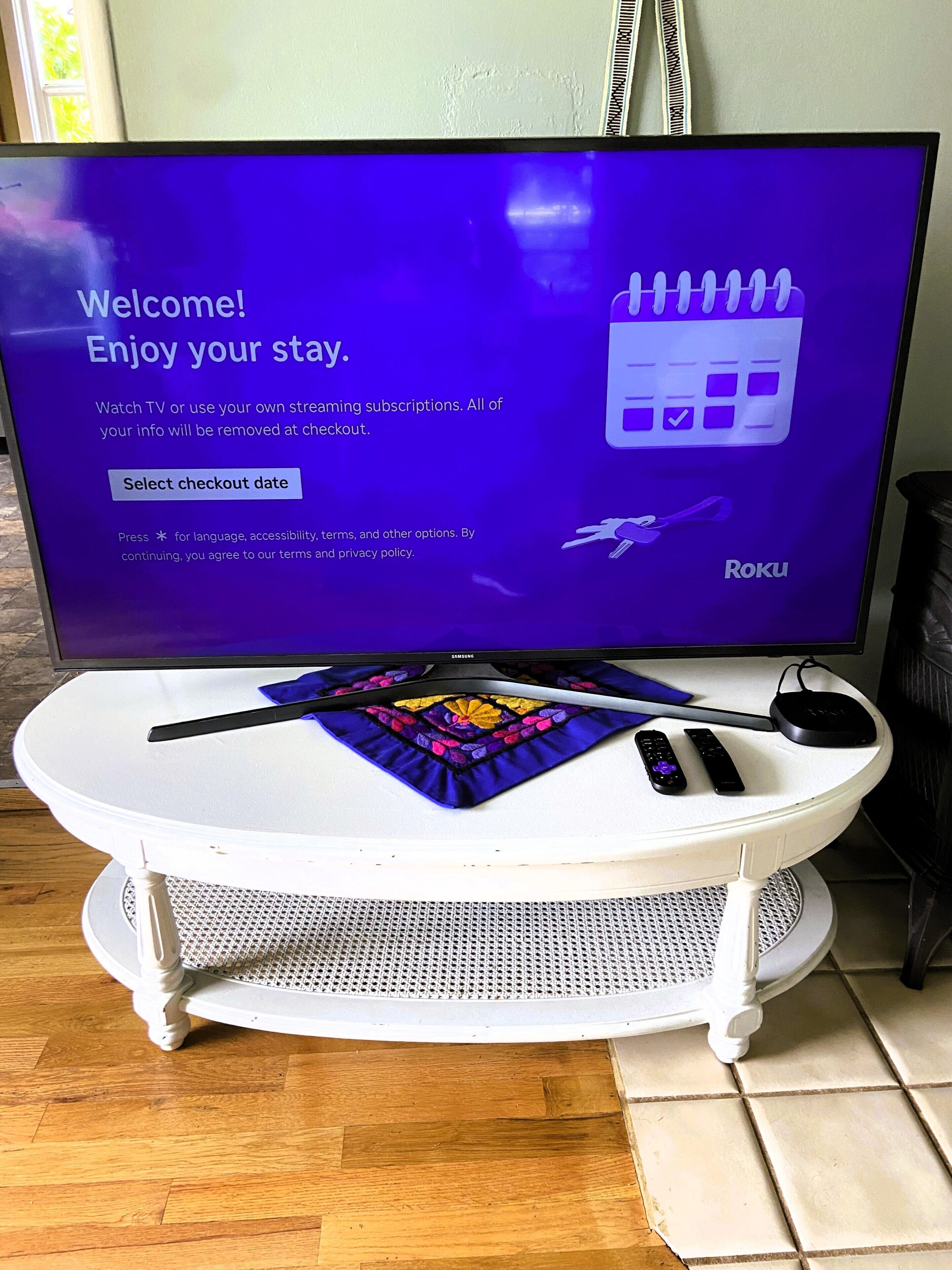 A television displaying a welcome message with a Roku logo, a remote control, and a smartphone on a white table with a decorative cloth.