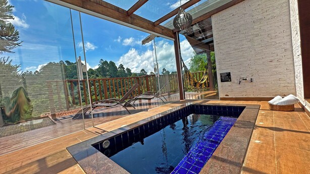 A glass-enclosed pool area with a view of a forest, wooden deck, and a white brick wall. - Estalagem da Montanha (Camanducaia)