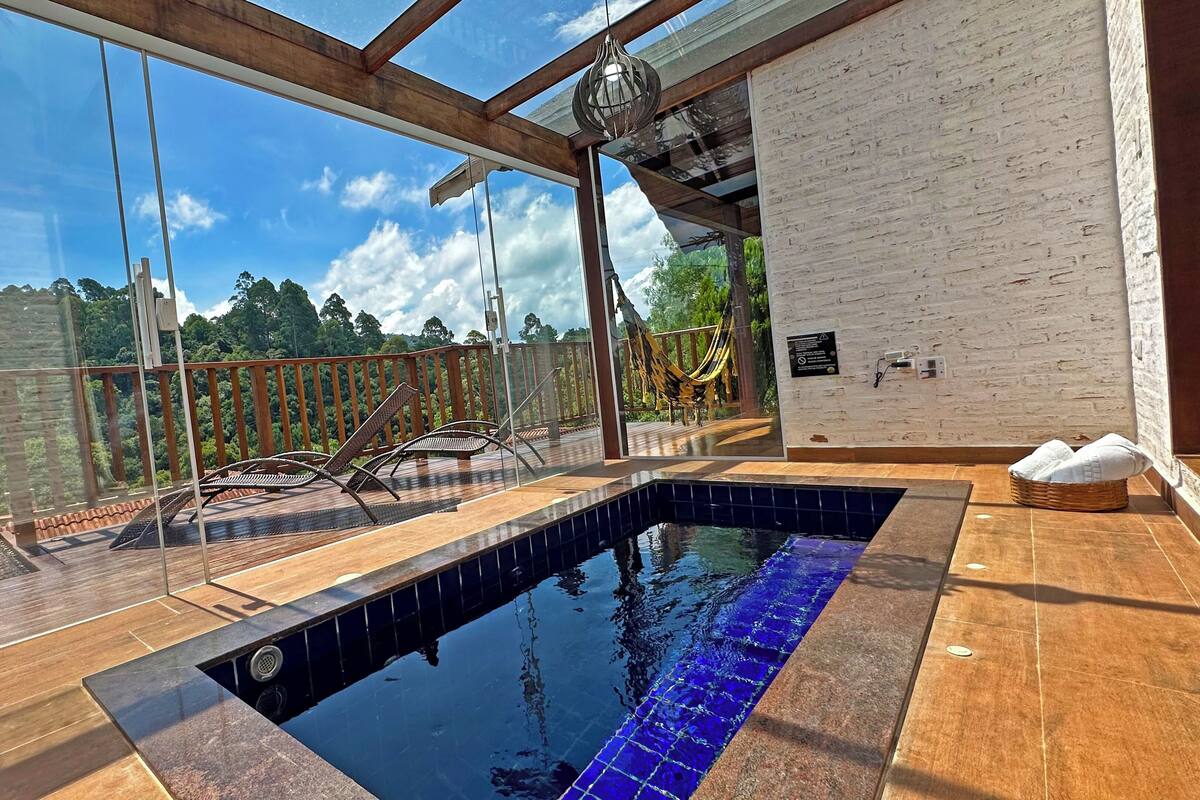 Un área de piscina con paredes de vidrio y vista a un bosque, terraza de madera y pared de ladrillo blanco.