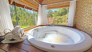 A hot tub with a wooden deck, a window with a view of greenery, and a towel on the deck. - Estalagem da Montanha (Camanducaia)