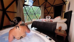 A room with a bed, a bathtub, and a window with a view of greenery.