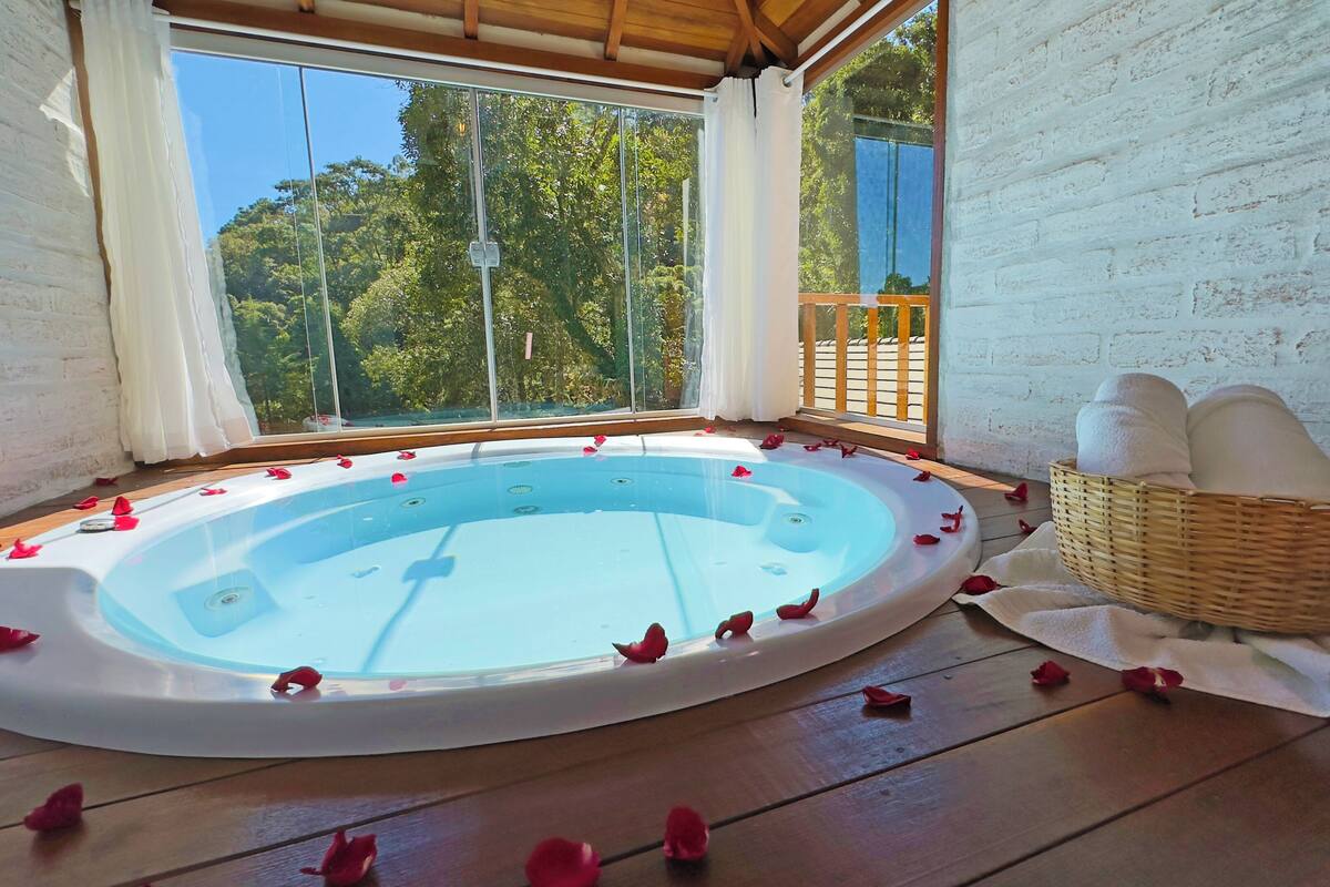 Un jacuzzi con pétalos de rosa en el suelo, una cesta de mimbre con toallas y vistas a árboles a través de grandes ventanales.