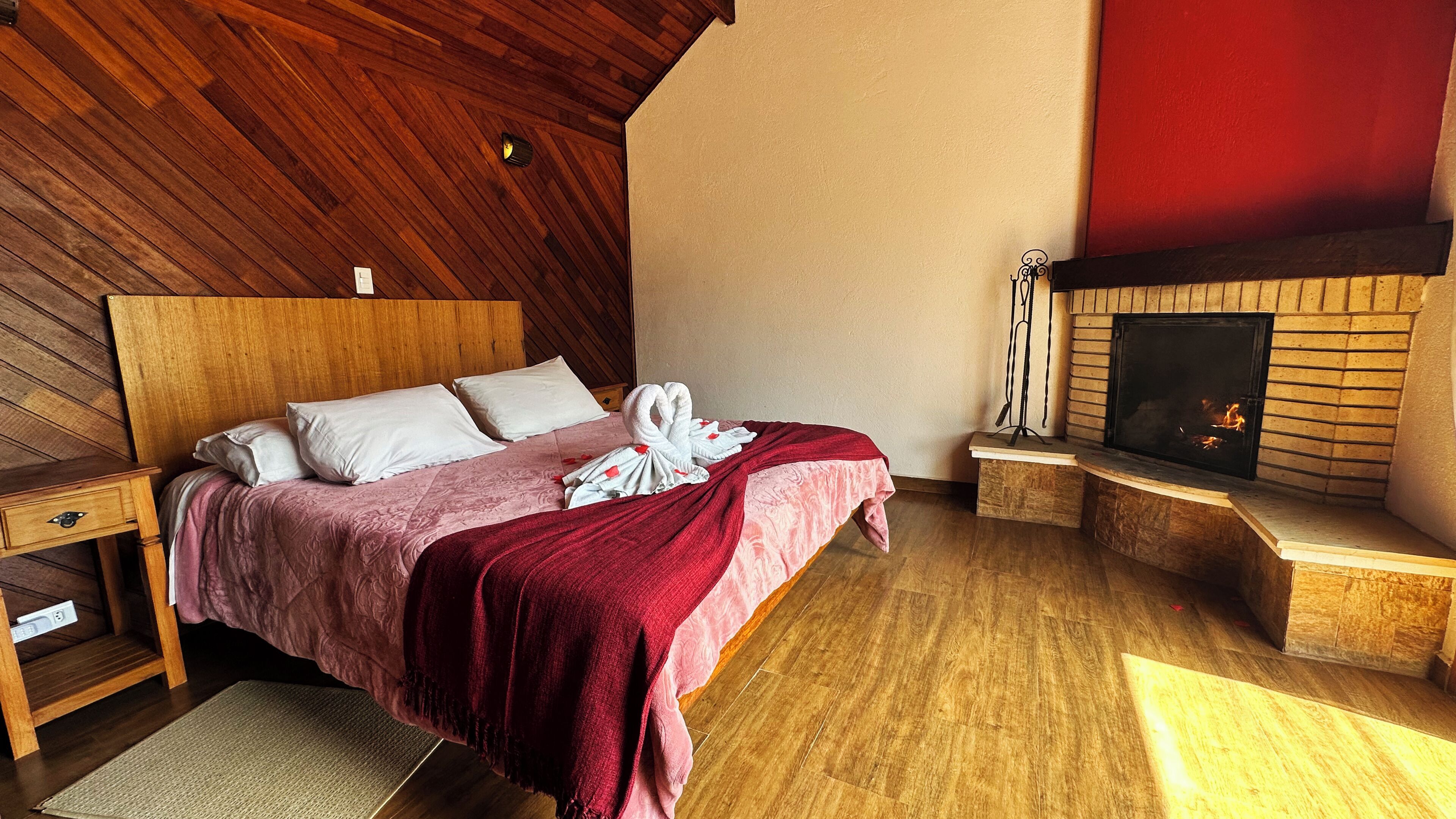 Una camera da letto con soffitto in legno, camino, un letto con coperta rossa e comodino.