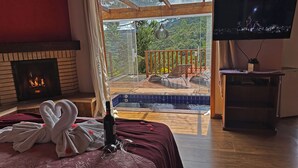 A room with a fireplace, a bed with towels, a bottle of wine, a TV, and a view of a pool and outdoor area. - Estalagem da Montanha (Camanducaia)