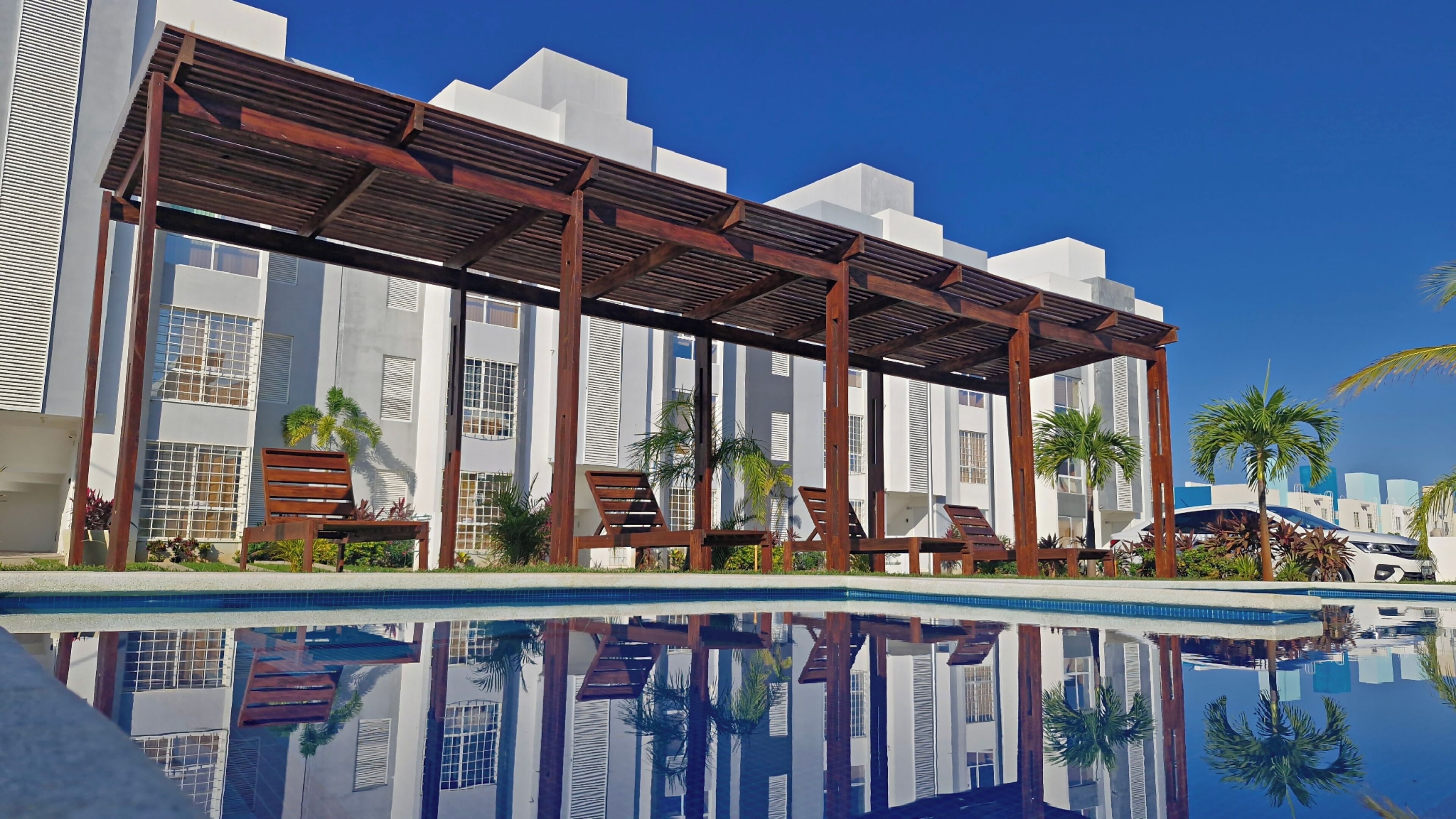 A modern hotel with a pool, wooden deck chairs, and palm trees.
