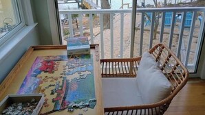 A room with a large window overlooking a dock and water. There is a wicker chair with a cushion and a partially completed puzzle on a table.