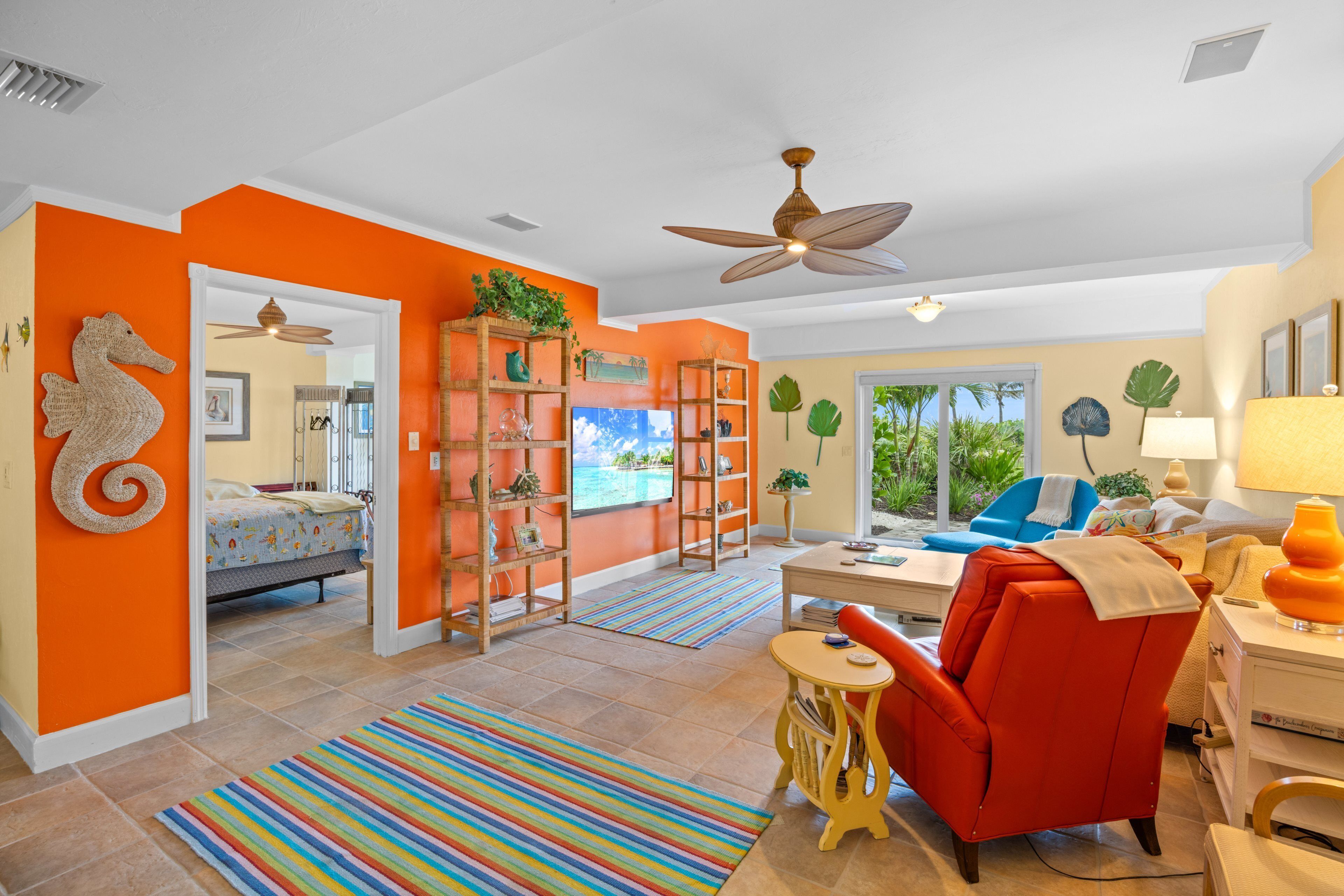 Un salon lumineux aux murs orange, avec un fauteuil rouge, une chaise bleue et une vue sur l’extérieur.