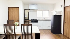 A compact kitchen with white cabinets, a black refrigerator, a stove, and a microwave. There is a dining table with two chairs and a vase of flowers. - 四葉草Clover House – Spacious Osaka Stay for Families & Groups (Osaka)