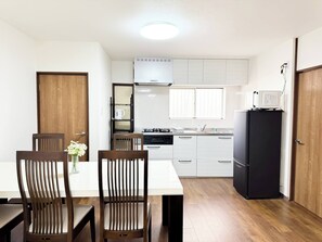 A compact kitchen with white cabinets, a black refrigerator, a stove, and a microwave. There is a dining table with two chairs and a vase of flowers. - 四葉草Clover House – Spacious Osaka Stay for Families & Groups (Osaka)